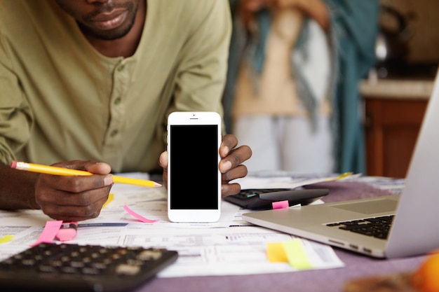 Cropped shot of African-American man holding mobile phone and pointing pencil at its blank screen