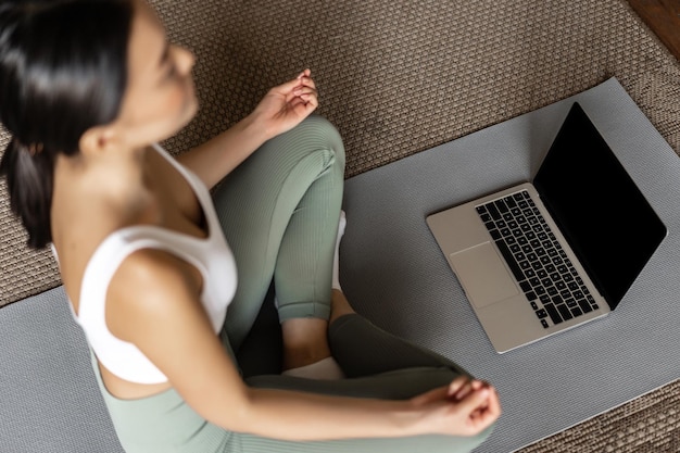 Cropped shot of asian woman meditating in floor mat at home listening to meditation podcast on lapto...