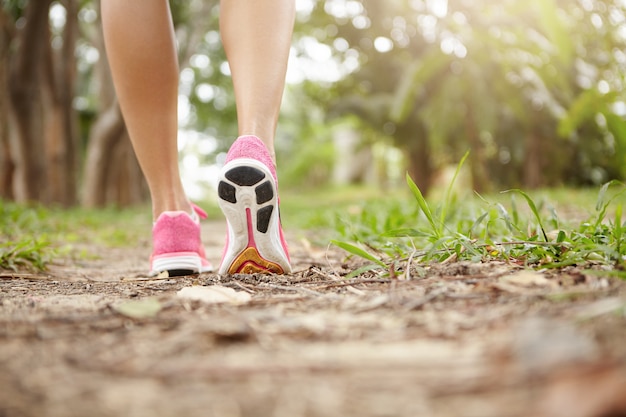 Cropped shot of athlete girl in pink running shoes hiking in forest on sunny day. Fit slim legs of sporty female in sneakers during jogging workout. Selective focus on sole.