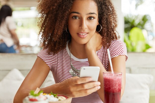 Cropped shot of attractive mixed race female model with bushy Afro hairstyle updates profile in social networks on mobile phone, connected to wireless internet in coffee shop, drinks cocktail