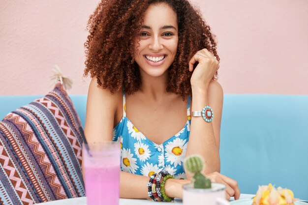 Cropped shot of beautiful African American female with curly hair, broad smile, enjoys leisure time in cafeteria, surrounded with fresh summer drink