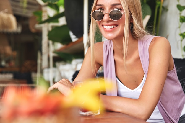 Обрезанный снимок красивой улыбающейся женщины в солнцезащитных очках и модной одежде, отдыхает в одиночестве в кафетерии с вкусным напитком, имеет счастливое выражение. Летом расслабленная женщина отдыхает в уютном баре