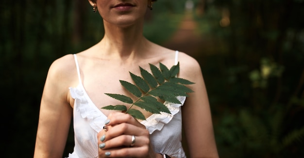 Обрезанный снимок красивой нежной молодой невесты в романтическом белом платье позирует на фоне зеленого леса, держа на груди лист папоротника. До неузнаваемости женщина отдыхает на открытом воздухе среди растений