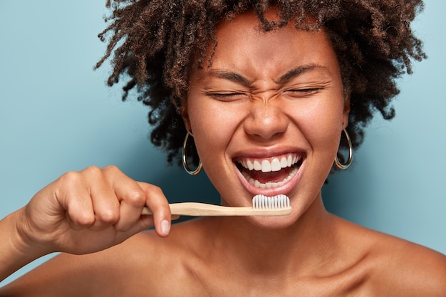 Cropped shot of cheerful glad dark skinned lady shows white teeth, has overjoyed expression, good mood in morning, prepares for dentist visit, stands with half naked body, isolated on blue wall