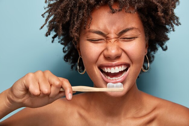 Cropped shot of cheerful glad dark skinned lady shows white teeth, has overjoyed expression, good mood in morning, prepares for dentist visit, stands with half naked body, isolated on blue wall