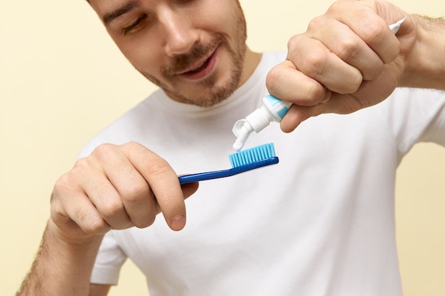 朝早く起きた後、歯ブラシにペーストを絞り、歯を磨く白いTシャツを着た元気で健康な若い白人男性のクロップドショット。