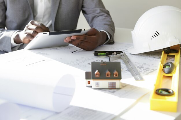 Cropped shot of dark-skinned contractor holding touch pad, entering data while working on new housing project, sitting at desk with drawings, scale model house, blueprint rolls, ruler and helmets