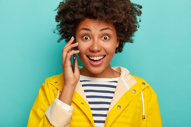 Cropped shot of dark skinned surprised young woman uses a mobile dressed in striped jumper and yellow raincoat