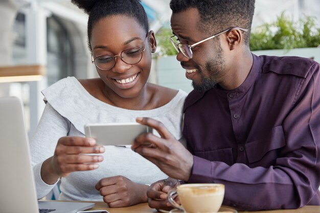 Cropped shot of deighted happy African couple holds smart phone horizontally, watch interesting video, have coffee break, smile joyfully, wears round spectacles.