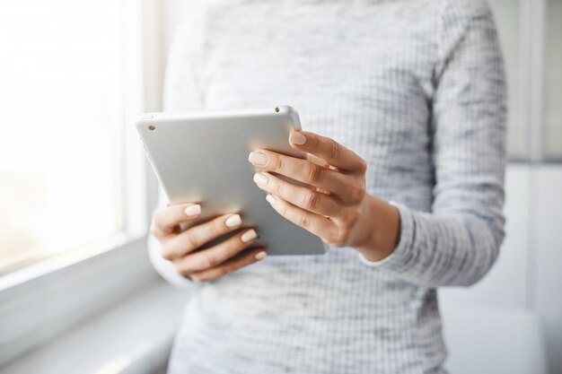 Cropped shot of employer standing near window, holding digital tablet, reading news on social media, checking her mail box, being busy while at work. Woman wants to take picture of beautiful scenery