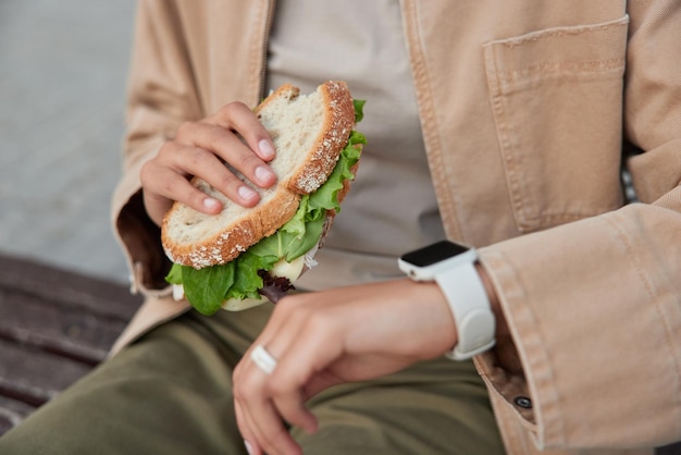 Cropped shot of faceless peson checks time on digital smartwatch eats delicious sandwich has snack rests after walking sits on bench checks covered distance. Street food and nutrition concept.