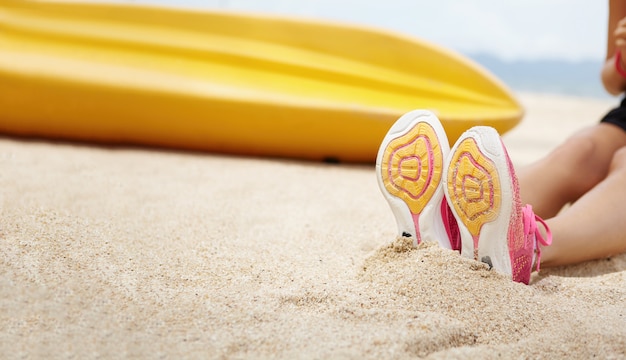 Foto gratuita colpo potato dell'atleta femminile che indossa le scarpe da corsa rosa che si siedono sulla spiaggia sabbiosa dopo l'esercizio attivo alla spiaggia. pareggiatore della donna che si rilassa all'aperto durante l'allenamento di mattina. messa a fuoco selettiva su suole