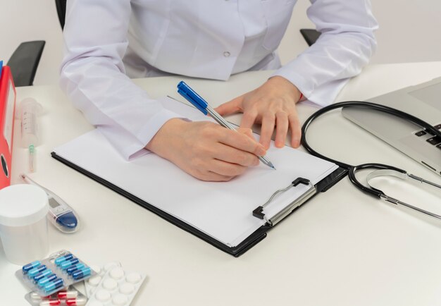 Cropped shot of female doctor hands working at desk with medical tools and laptop writing prescription on clipboard