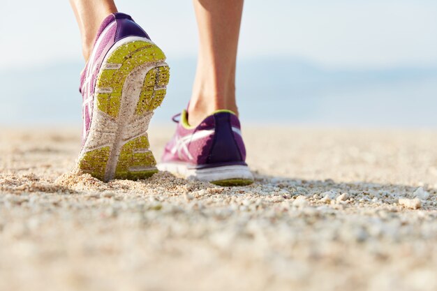 Cropped shot of female`s legs in purple training shoes stands at sandy beach on coastline