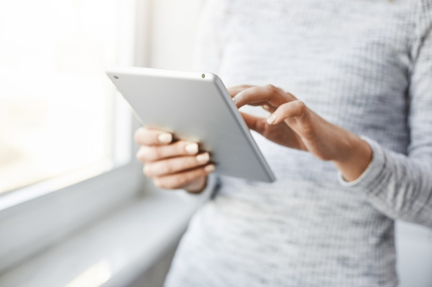 Cropped shot of female student standing near window, touching screen of digital tablet, searching in web or communicating with peers while sitting in home ill. Girl edits her post on page in her blog