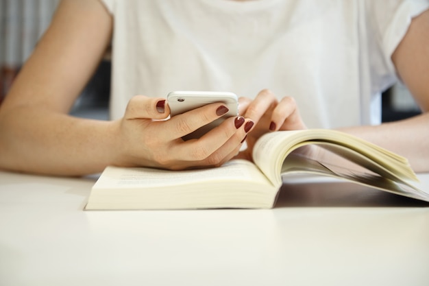 Cropped shot of female with neat manicure wearing white blouse with her hands on an open book, browsing the Internet using mobile phone while studying and searching for information at the library