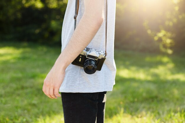 Cropped shot of male`s body with retro camera on shoulder standing at green grass going to photographing someone