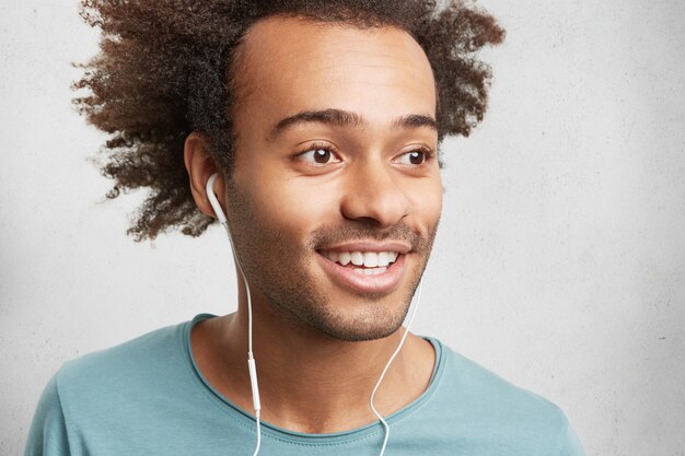 Cropped shot of mixed race unshaven man looks happily as listens to pleasant songs in earphones