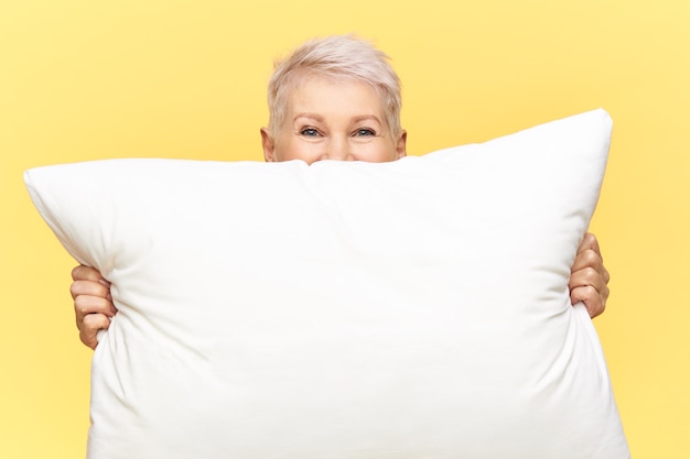 Обрезанный снимок красивой женщины средних лет с короткими волосами, прячущейся за белой большой перьевой подушкой с копией пространства
