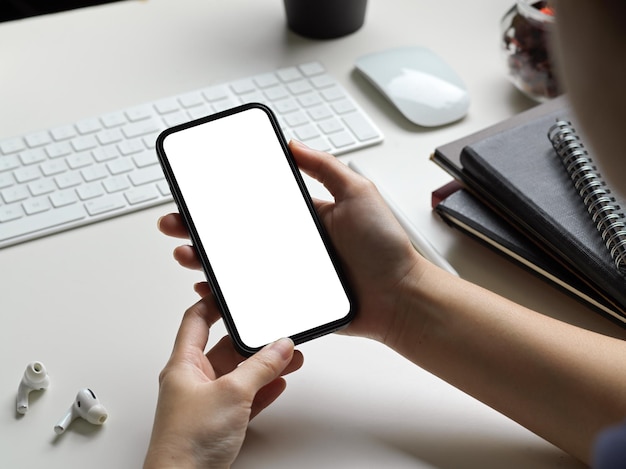 モダンな白いオフィスのワーキングデスクで空白の画面のスマートフォンを保持している女性または女性のトリミングされたショット