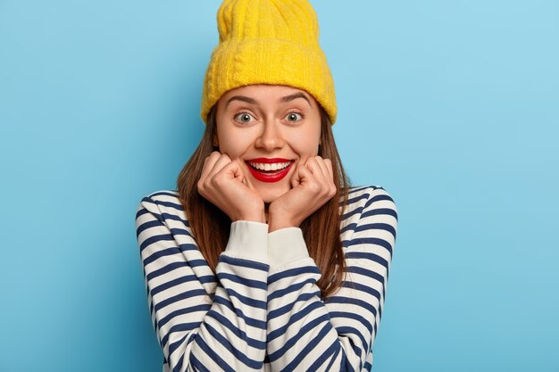 Cropped shot of pleasant looking young woman smiles broadly, has white teeth, wears red lipstick, keeps hands under chin, wears yellow hat, striped sailor jumper, grins joyfully, has awesome day