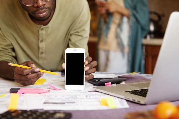 Cropped shot of serious African American man in glasses pointing pencil at blank screen mobile phone