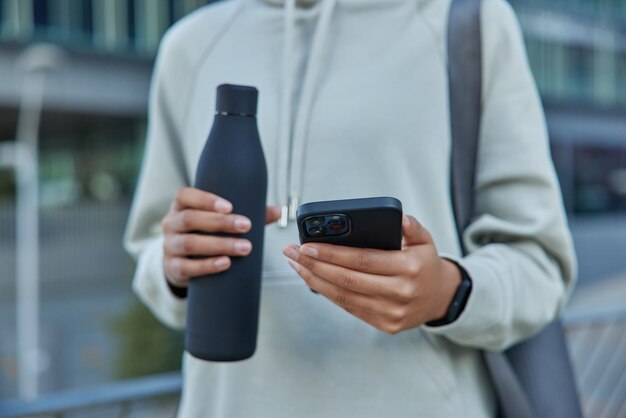 Cropped shot of sportswoman in casual hoodie holds bottle of water and mobile phone checks fitness activity carries rolled mat poses outdoors