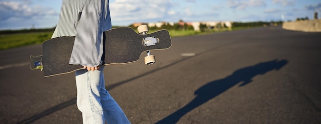 コンクリートの道路でスケートボードで歩いているロングボードを握っているティーンスケーターの女の子の手をカットした写真