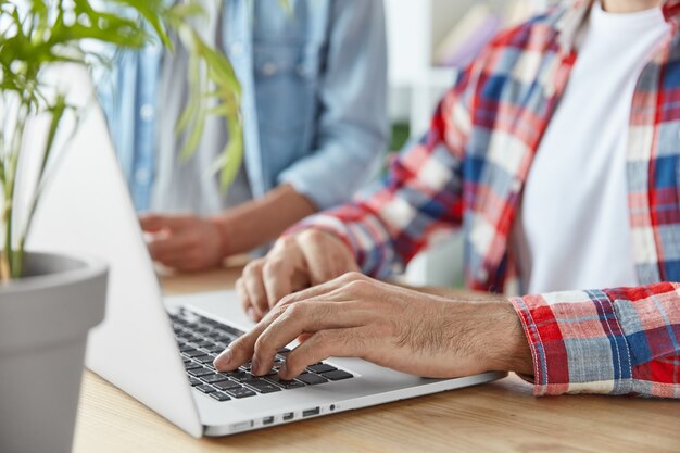 Cropped shot of two male bloggers type publication on laptop computer, use laptop computer, sit at wooden desk. Young prosperous bussinessmen check mail and send feedbacks, connected to wifi