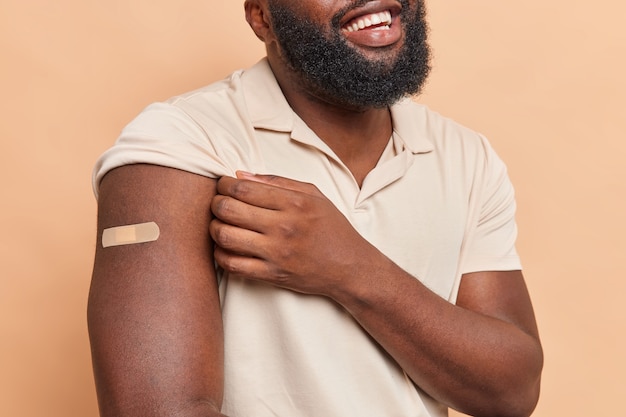 Free photo cropped shot of unrecognizable man with thick beard shows arm with plaster glad to be vaccineted takes care of health dressed in casual t shirt isolated over brown wall