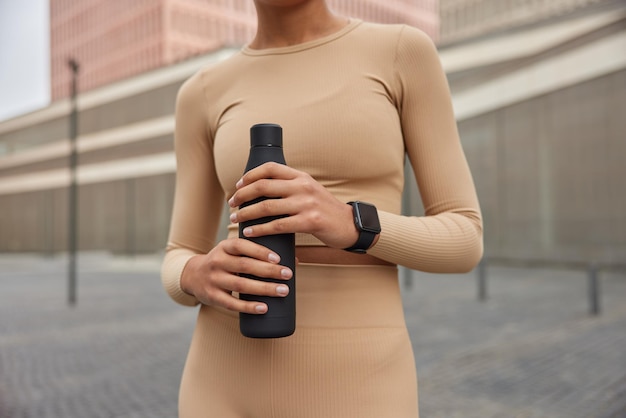Обрезанный снимок неузнаваемой женщины, одетой в спортивную одежду, держит спортивную бутылку пресной воды, носит смарт-часы, чувствует жажду после кардиотренировок, стоит на открытом воздухе на размытом фоне
