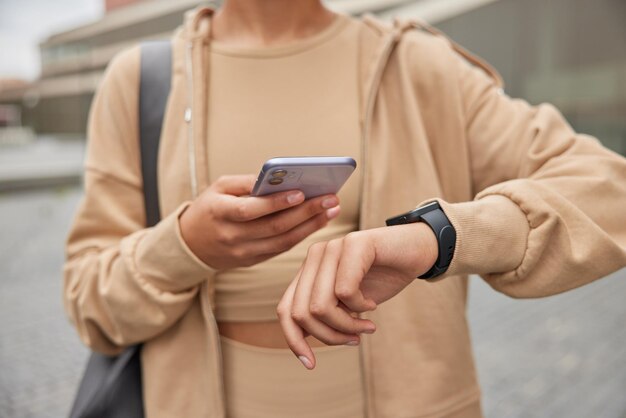 Cropped shot of unrecognizable woman holds modern mobile phone checks results of fitness traning dressed in sportswear carries fitness mat poses outdoors. People sport and technology concept