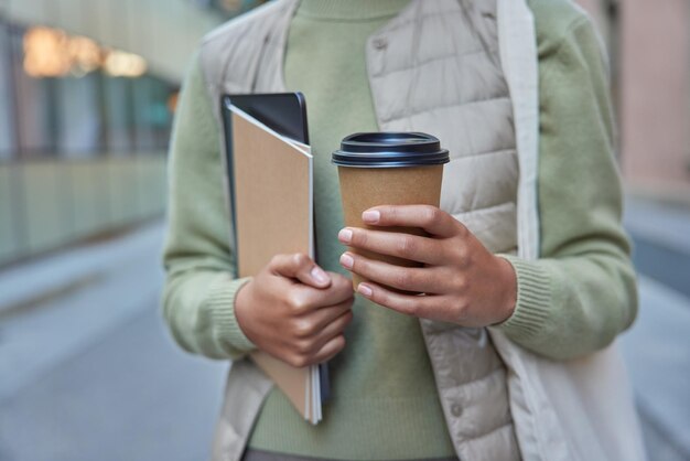 Cropped shot of unrecognizable woman holds takeaway aromatic coffee notebook and digital tablet dressed in casual clothes strolls outdoors during daytime Faceless female model goes somewhere alone