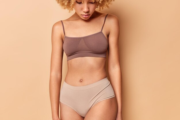 Cropped shot of unrecognizable woman wears top and panties has perfect body shape shows abdomen after liposuction isolated over beige background. Ideal figure and slimming concept. Torso of fit female