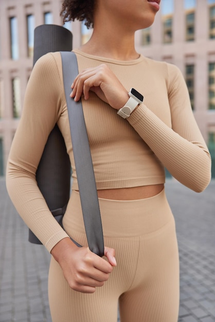 Cropped shot of unrecognizable woman with perfect figure dressed in activewear wears smartwatch carries rolled karemat going to have fitness training exercises outdoors enjoys healthy lifestyle
