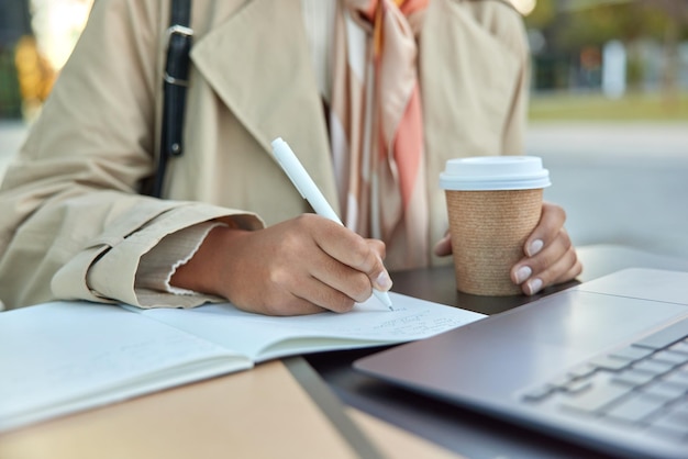 Cropped shot of unrecognizable woman works distantly sits at desktop in front of laptop computer makes notes in diary drinks takeaway coffee Unknown businesswoman plans strategy prepares project