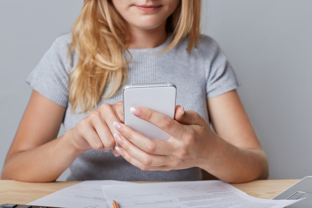 Cropped shot view of blonde female enterpreneur holds smart phone, surrounded with documents, recieves messages