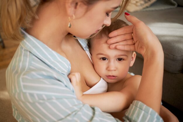 Cropped shot of worried young woman holding her sad infant son, checking if he has fever without thermometer, feeling forehead with back of hand. Childhood, sickness, health and motherhood concept