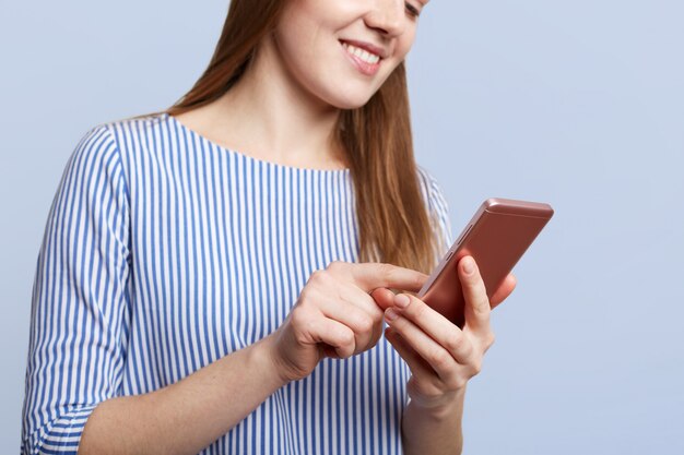 Cropped shot of young female with cheerful expression holds modern cell phone, messages with friends, connected to wireless internet