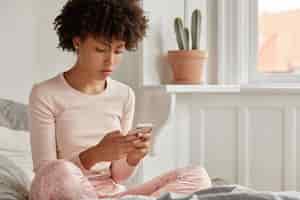 Free photo cropped shot of young lady blogger with afro haircut, dressed in pyjamas, holds smart phone