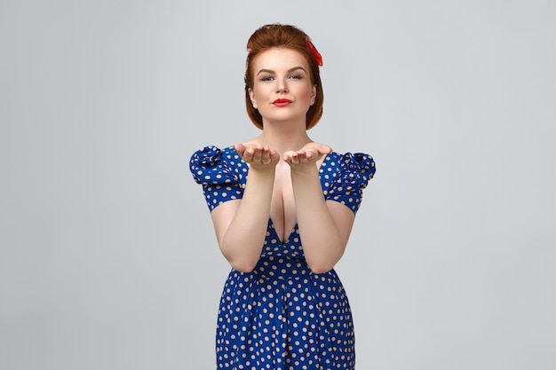 Cropped studio shot of attractive stylish young caucasian female wearing bright make up and retro blue dotted dress with low cut neck holding open palms and pouting lips, blowing air kisses