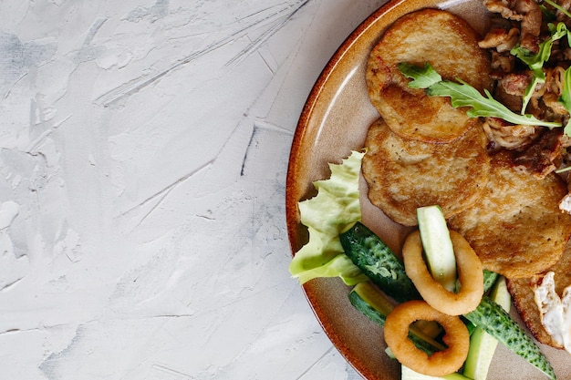 Free photo cropped topview of tasty potato puncakes breakfast served with fried eggs fresh green cucumbers fried golden onion and crusty meat served with delicious salad leaves luying on brown clay plate