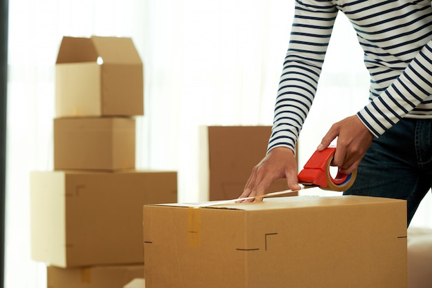 Free photo cropped unrecognizable man applying adhesive tape to the box with dispenser