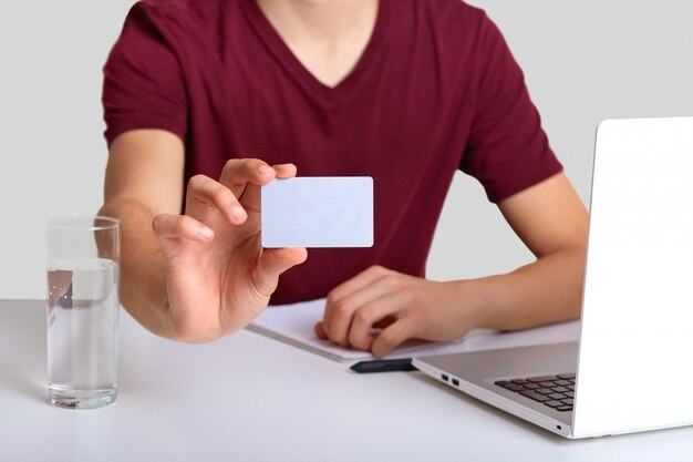 Cropped unrecognizable man in casual red t shirt, sits at workplace with laptop computer, glass of water, focus on blank card with free space for your advertising content or promotion