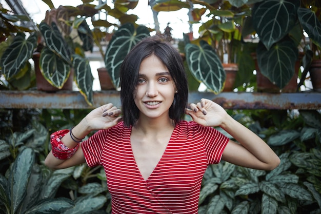 Cropped view of beautiful young European female with dark bib hairstyle relaxing outdoors, walking in jungle surrounded with exotic plants, feeling happy and carefree, smiling joyfully at camera