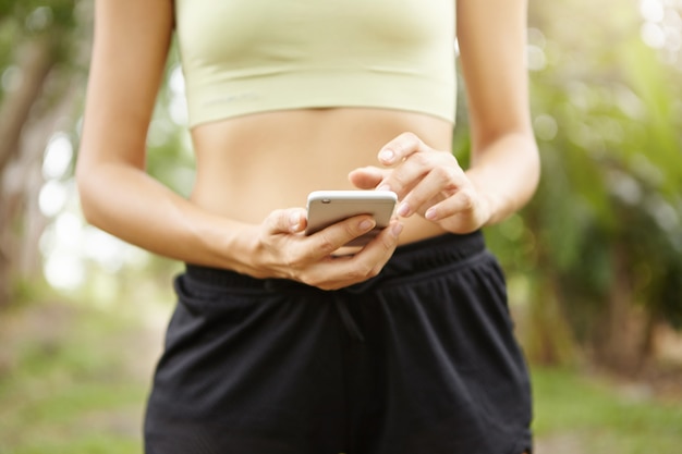 Cropped view of fit female jogger in sports bra and black shorts using her mobile phone before run.