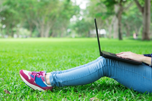 Vista ritagliata della ragazza che si siede sull'erba con il computer portatile