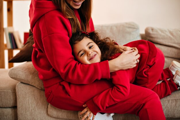 Cropped view of laughing mom embracing daughter at home. Indoor shot of laughing kid having fun with mother.