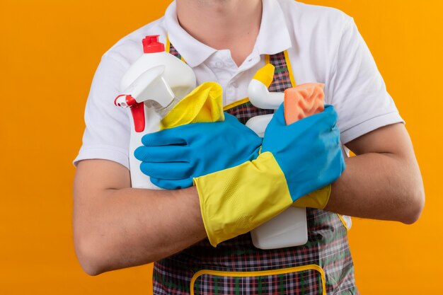 Cropped view of man wearing apron and rubber gloves holding cleaning supplies and sponge over isolated orange background