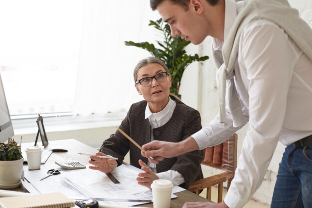 Cropped view of talented ambitious young man architect holding pencil and pointing at drawings on desk while showing construction project to his senior woman boss in glasses. Working in team